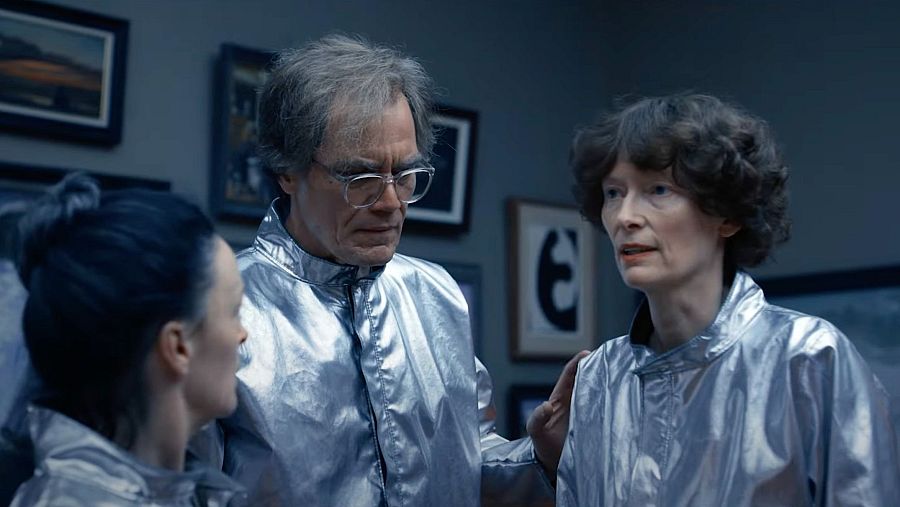 Tres personas en chaquetas plateadas conversan en una habitación con iluminación azulada.  Una mujer toca el hombro de un hombre que mira a otra mujer.