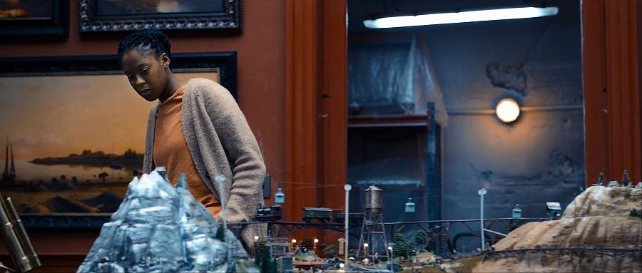 Mujer con trenzas observa maqueta detallada de paisaje montañoso con tren miniatura; viste camiseta naranja y chaqueta beige; habitación con paredes rojas y cuadros.