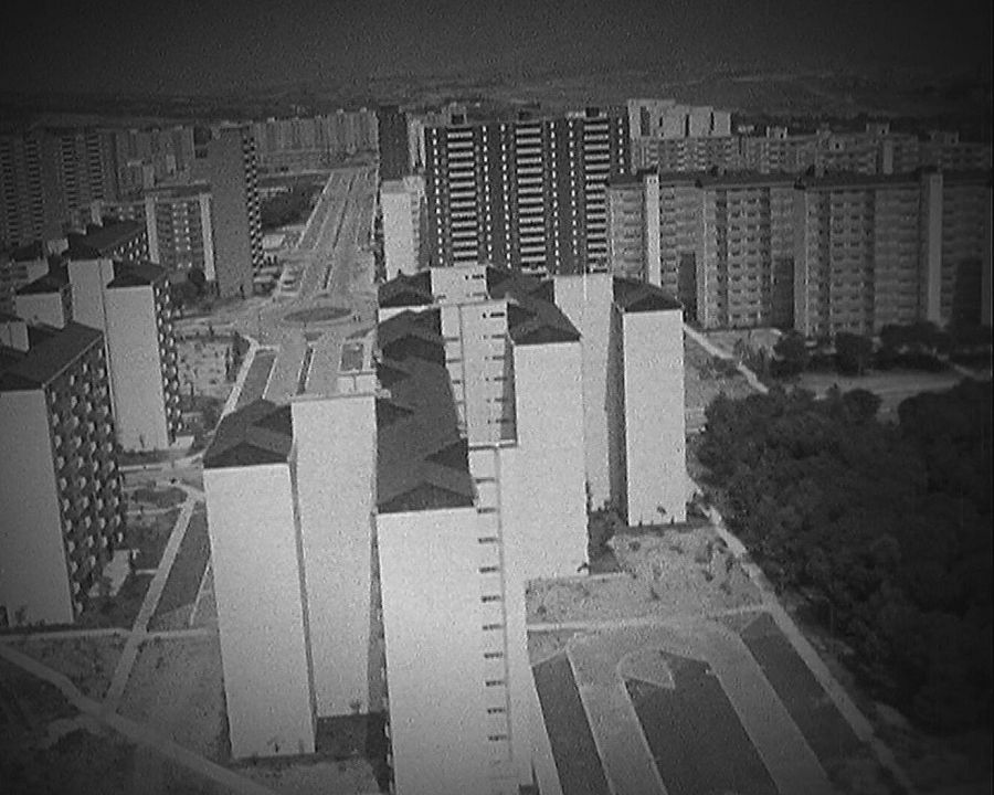 Fotografía aérea en blanco y negro de bloques de apartamentos de varios pisos, con calles, zonas verdes y una rotonda.  Aspecto funcional y ausencia de vehículos.