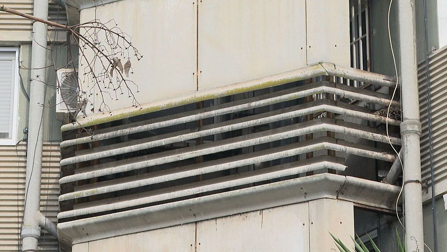 Conducto de evacuación de humos gris claro con rejillas horizontales, en fachada beige.  Se ven tuberías, cables y vegetación.