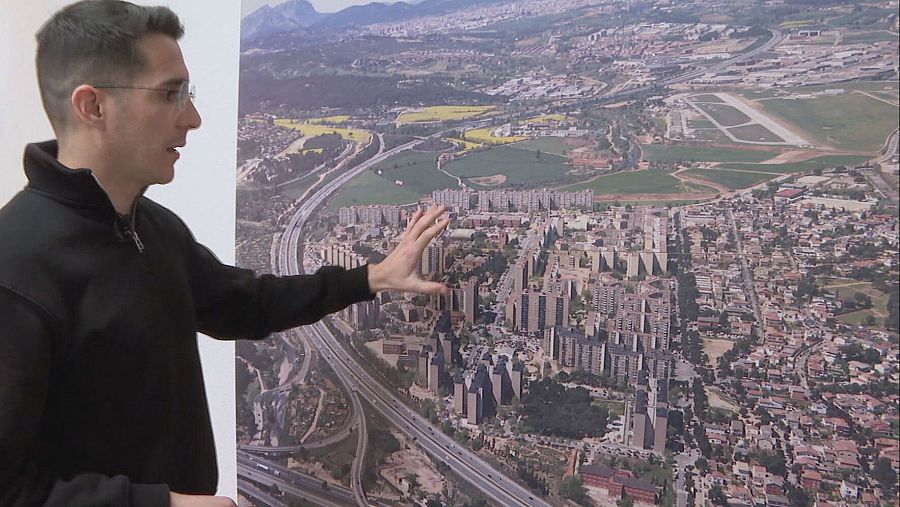 Alcalde de perfil, con gafas y sudadera oscura, señala un mapa aéreo que muestra una zona urbana con edificios, carreteras y espacios verdes.