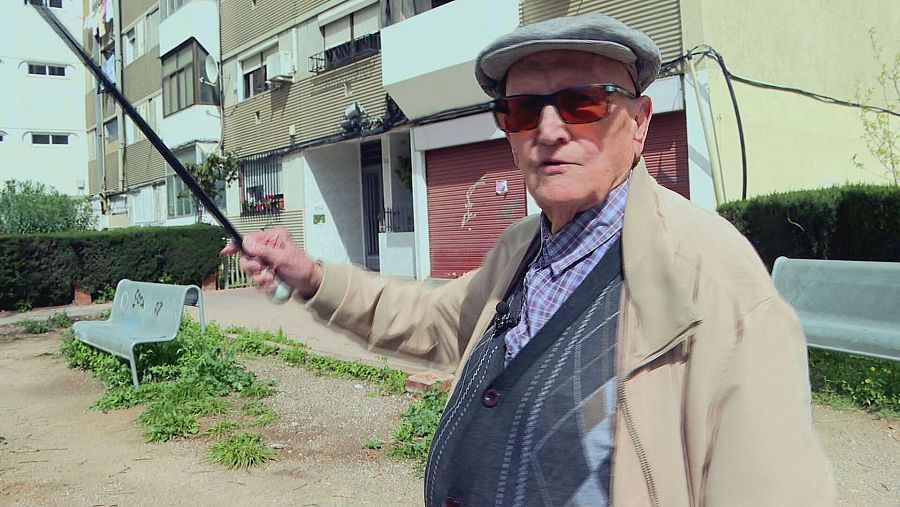 Hombre mayor con boina gris, gafas de sol y chaleco, gesticulando con un bastón y micrófono inalámbrico, en un entorno exterior con bancos y edificios al fondo.