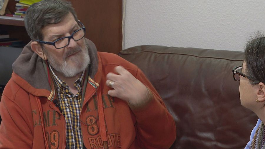 Entrevista a un hombre con sudadera naranja y gafas que gesticula mientras habla; la entrevistadora, con gafas y pañuelo, escucha atentamente.