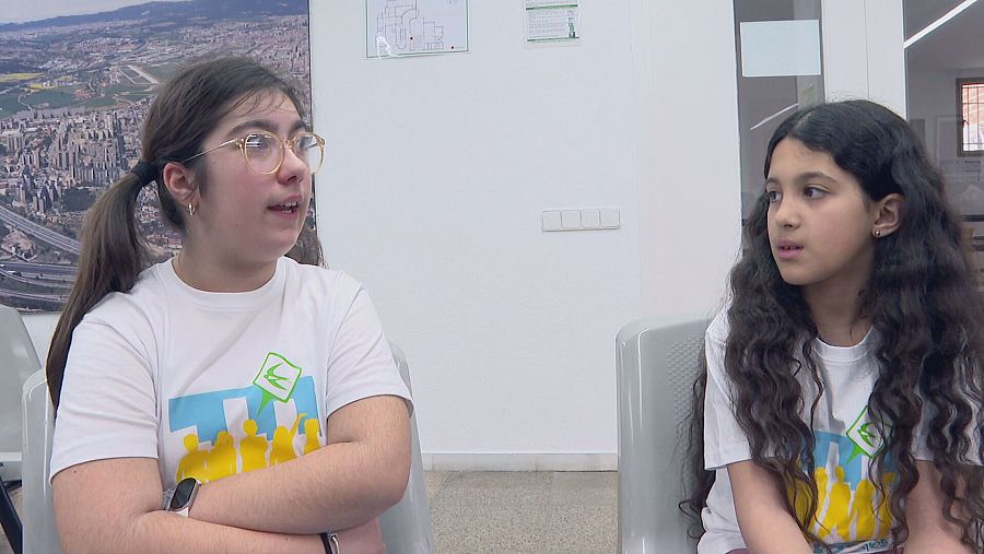 Dos niñas con camisetas blancas y logo amarillo participan en una reunión en una sala con imágenes en la pared. Una lleva el pelo recogido y gafas; la otra, el pelo largo y rizado.