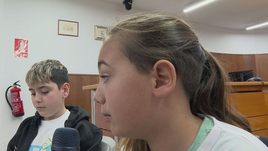 Niños en una reunión, con un micrófono delante. Él viste camiseta blanca y chaqueta oscura; ella, camiseta blanca con detalle verde en el cuello.