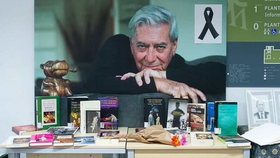 Altar conmemorativo con retrato de Vargas Llosa, libros, flores y escultura de hipopótamo en biblioteca. Cartel informativo de plantas visible.