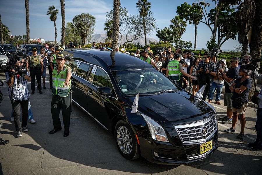 Coche fúnebre negro con matrícula AJR-827, rodeado de periodistas y seguridad.  Posible bandera peruana en el capó.  Entorno cálido con palmeras.