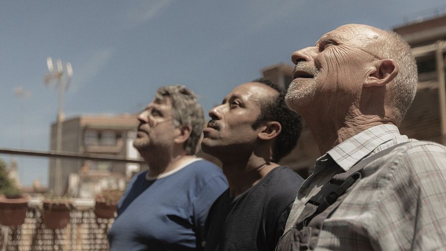 Tres hombres, con vestimentas variadas, miran hacia arriba en una escena que parece una azotea o terraza con edificios al fondo.