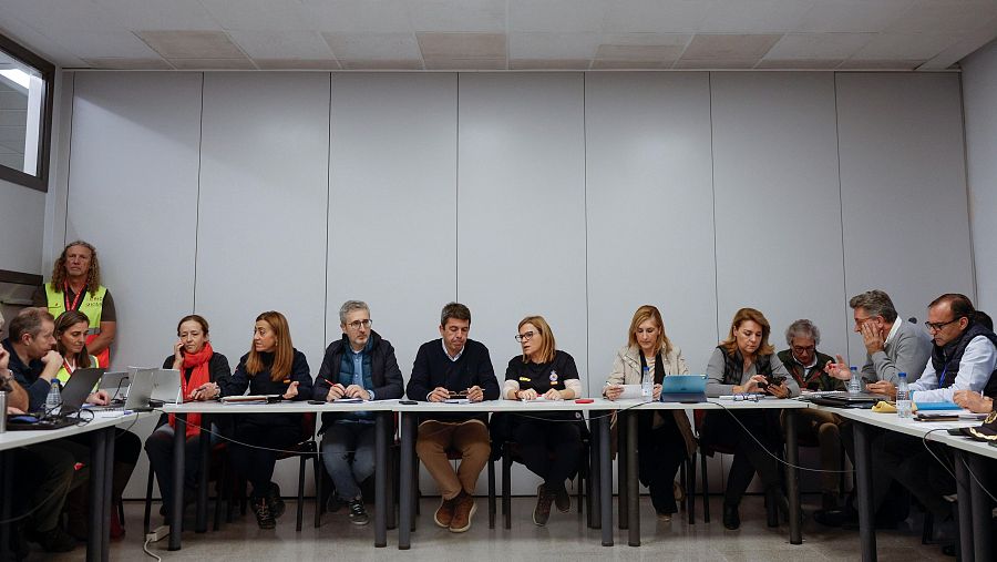 Personas reunidas alrededor de una mesa con ordenadores y documentos, trabajando en una situación de emergencia.  Ambiente serio y concentrado.