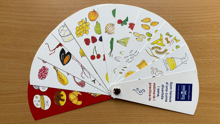 Abanico ilustrado con variedad de alimentos: frutas, verduras, carnes y mariscos.  Incluye el nombre 'Carme Ruscalleda' y la frase 'Som hereus d'una cuina divertida i sana, estima-la i practica-la'.