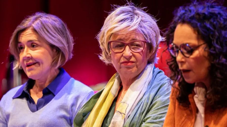 Tres mujeres en un evento cultural; una con jersey azul, otra con chaqueta verde y bufanda, y la tercera con chaqueta naranja. Fondo rojo.