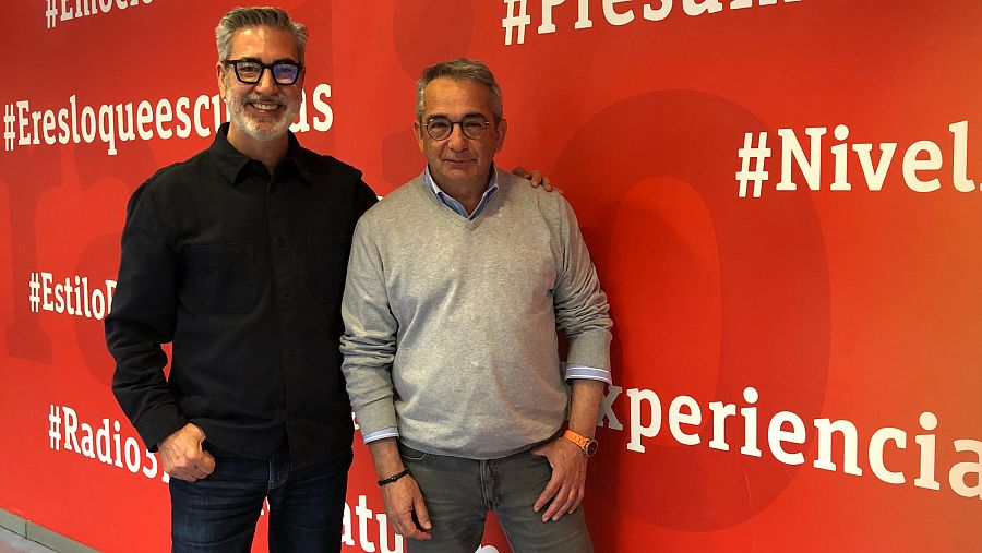 Dos hombres sonrientes posan ante un fondo rojo con hashtags y palabras en blanco. Uno viste camisa negra y el otro un jersey gris.