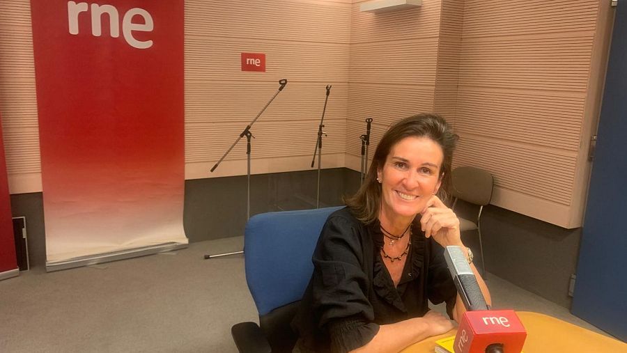 Presentadora de RNE Valladolid en estudio de radio, sonriendo a cámara con micrófono y logo RNE visible al fondo.