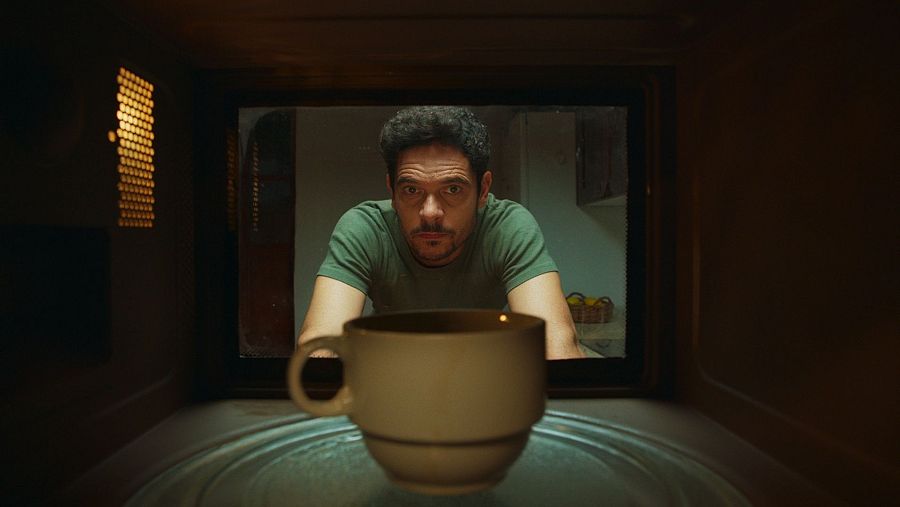 Un hombre observa una taza beige dentro de un microondas; pelo oscuro y rizado, barba corta, camiseta verde. Fondo: cocina desenfocada.
