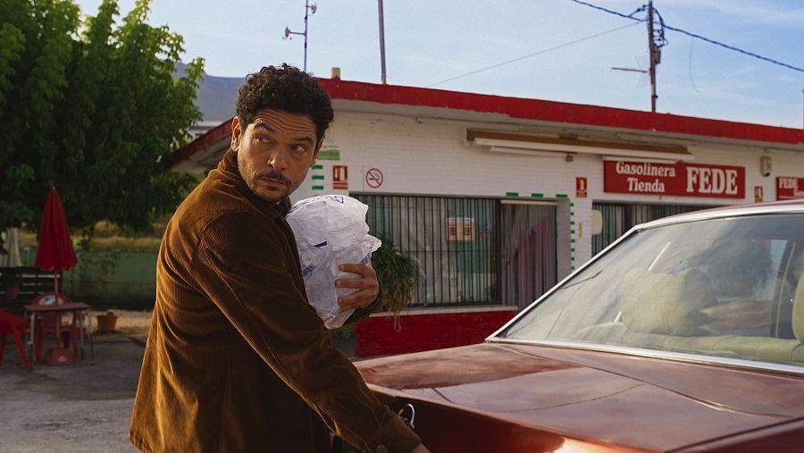 Hombre con chaqueta marrón carga hielo en gasolinera 'Tienda FEDE', con terraza roja cercana y coche granate aparcado.