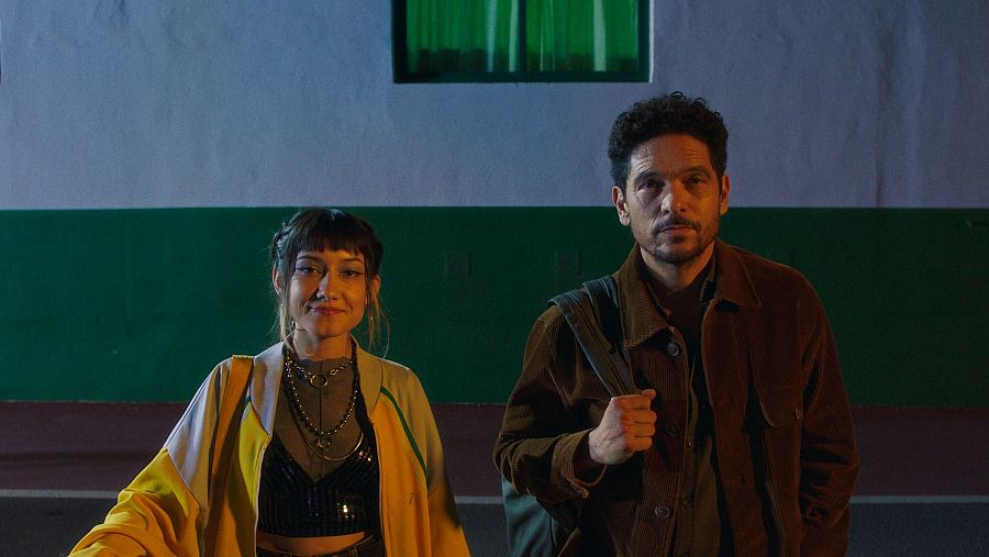 Dos personas, un hombre y una mujer, miran a cámara. Él viste chaqueta marrón y mochila; ella, chaqueta con paneles amarillos y negros y varios collares. Escena nocturna.