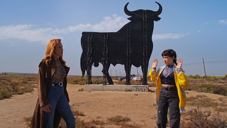 Dos mujeres, una con gabardina marrón y otra con chaqueta amarilla, aparecen en primer plano ante un toro de Osborne en un paisaje desértico.