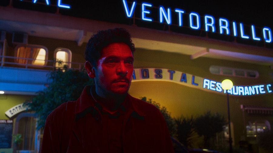 Hombre con chaqueta granate y expresión seria, frente a un edificio iluminado con letreros de 'VENTORRILLO' y 'HOSTAL RESTAURANTE'.