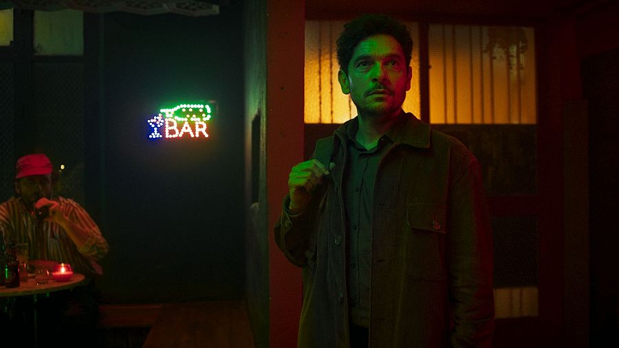 Intensa escena nocturna de un bar en 'Lo carga el diablo'. Un hombre en primer plano, con chaqueta oscura, mira con tensión. Al fondo, otro hombre con gorra roja bebe.