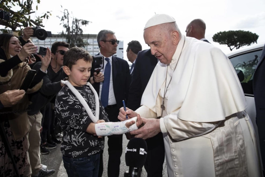 El Papa firma el yeso de un niño con un bolígrafo; el niño viste un jersey oscuro; la escena es documentada por varios presentes.