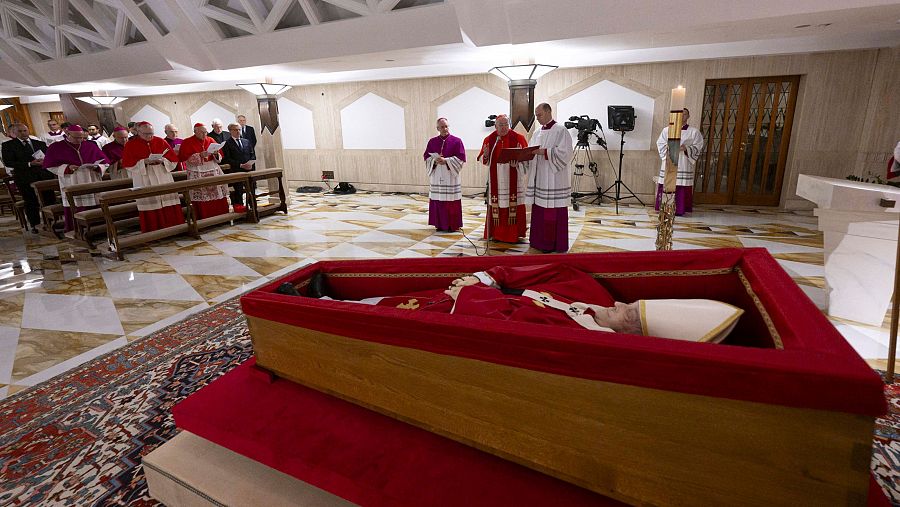 Féretro de madera con forro rojo en capilla, rodeado de cardenales y clérigos vestidos de rojo y blanco.  Equipos de grabación presentes.