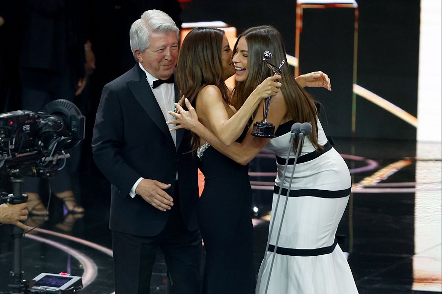 Sofía Vergara recibe un premio, abrazada por Eva Longoria.  Un hombre en esmoquin está cerca.  Escena de una ceremonia.