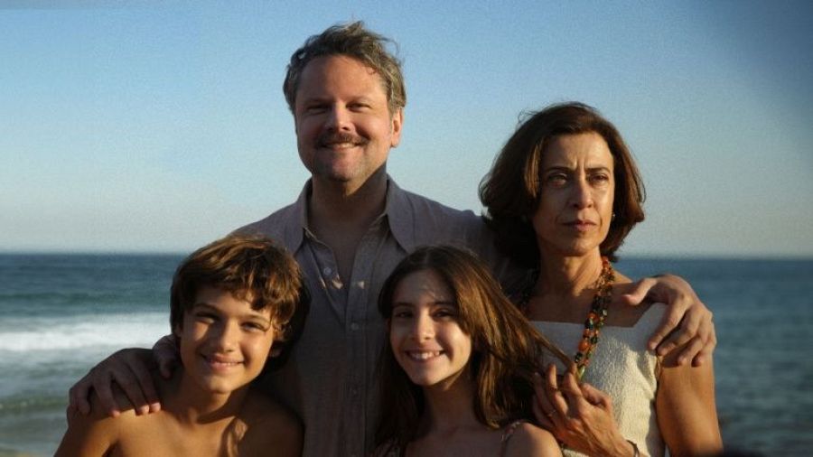 Familia de cuatro en playa soleada; padre con camisa gris abraza a sus hijos, madre con vestido claro y cabello castaño apoya su mano en su hija.