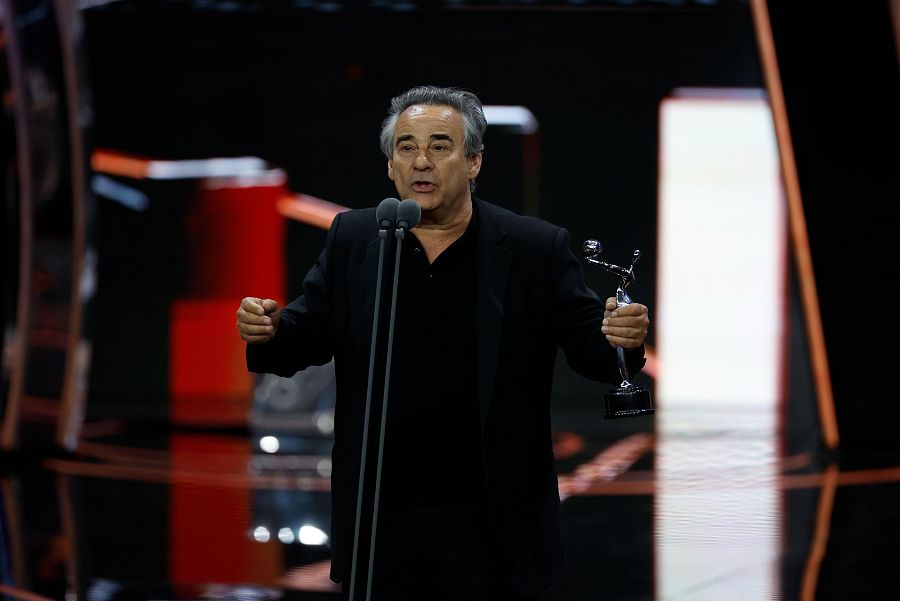 Actor con chaqueta negra da un discurso, sosteniendo un premio y hablando por micrófono. Fondo oscuro con reflejos rojos.