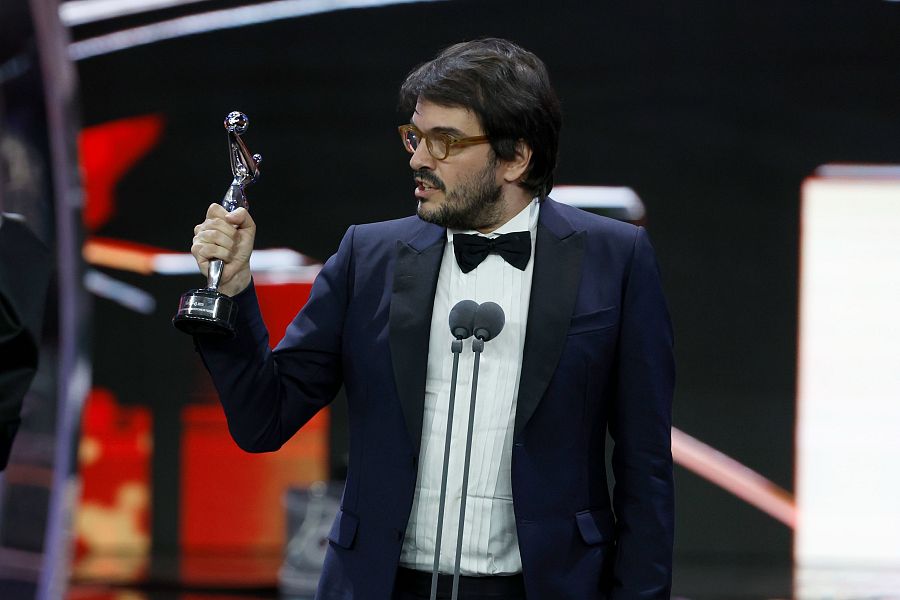 Productor elegante en traje azul marino y pajarita recibe premio, sosteniendo una estatuilla plateada ante dos micrófonos.