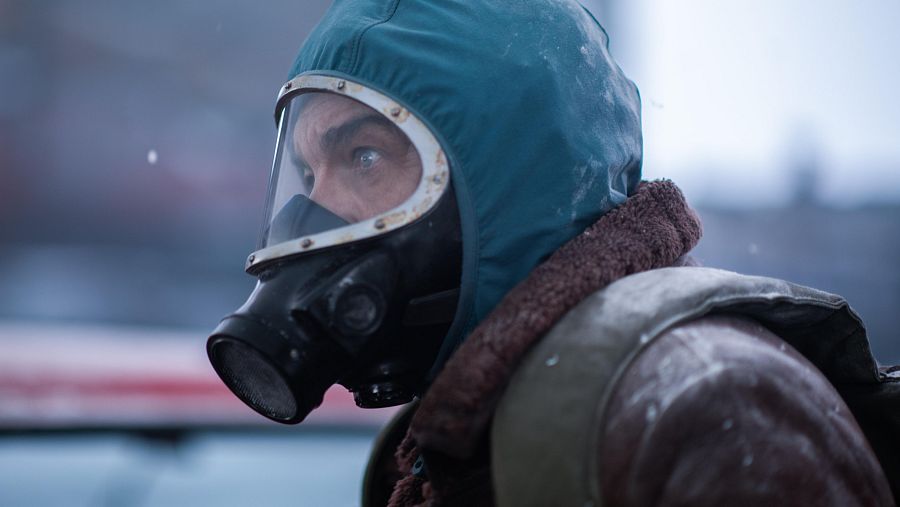 Ricardo Darín, con máscara antigas negra y capucha verde, viste ropa de abrigo marrón en una escena que sugiere un ambiente frío y peligroso.