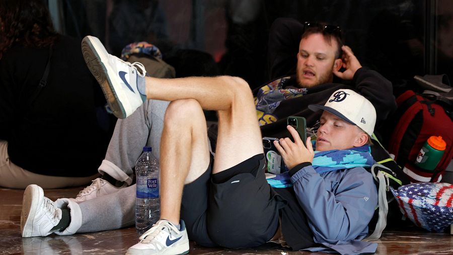 Pasajeros esperan en una estación, sentados en el suelo con mochilas y una botella de agua. Uno lleva pantalones cortos y zapatillas; otro, gorra y sudadera.