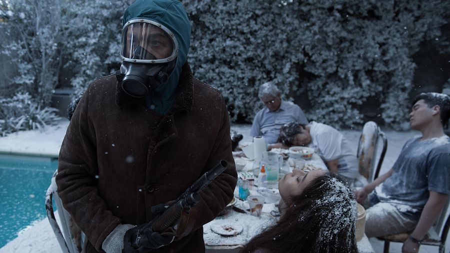 Escena nevada con mesa, restos de comida y varias personas. Una figura con traje protector y arma larga observa el entorno. Atmósfera desoladora.