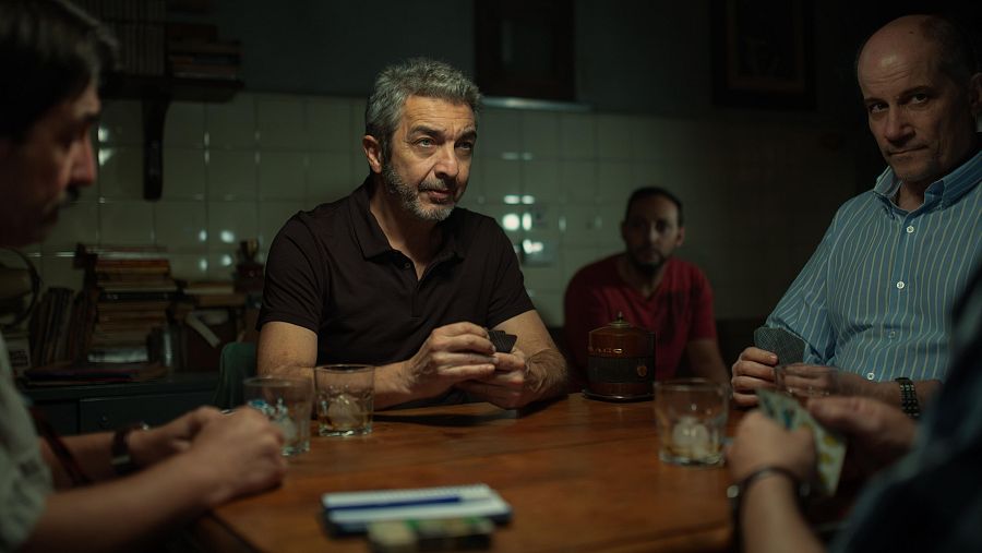 Cuatro hombres juegan a las cartas en una habitación oscura; tensión palpable, vasos y tabaco sobre la mesa de madera.