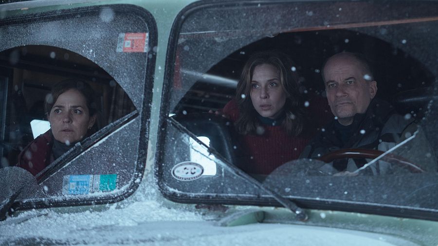 Tres personas en un vehículo antiguo, con parabrisas roto, observan algo con preocupación durante una nevada.  Se ve una pegatina en el parabrisas.
