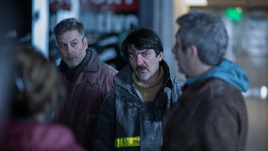 Escena de 'El Eternauta': cuatro personas en un espacio interior oscuro; tres hombres conversan, uno con una insignia 