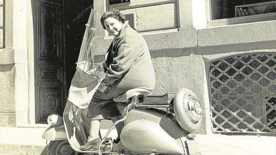 Gloria Fuertes sonríe a cámara sentada en una Vespa clara con rueda de repuesto; imagen en blanco y negro, abrigo oscuro.
