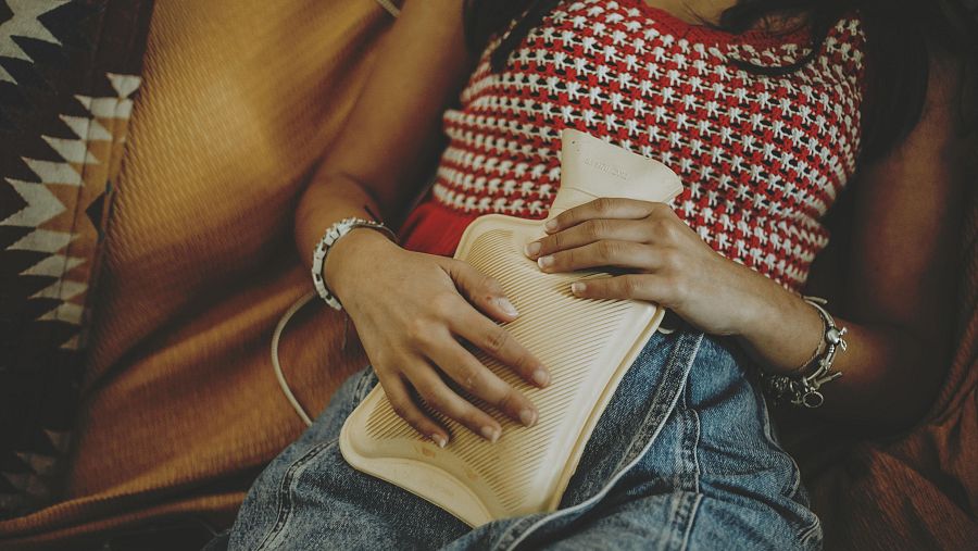 Mujer en sofá con camiseta a cuadros rojos y blancos, vaqueros y pulseras, usando una bolsa de agua caliente para aliviar el dolor abdominal.