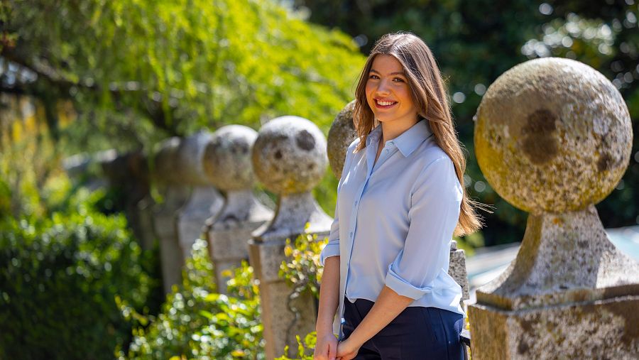 Infanta Sofía, sonriente, en jardines. Camisa azul claro, pantalones oscuros, cabello suelto. Apoyada en balaustrada de piedra.
