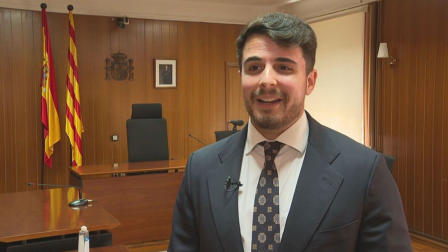 Hombre en traje oscuro con corbata geométrica, en sala con paneles de madera y banderas, frente a un micrófono en una mesa. Contexto formal.