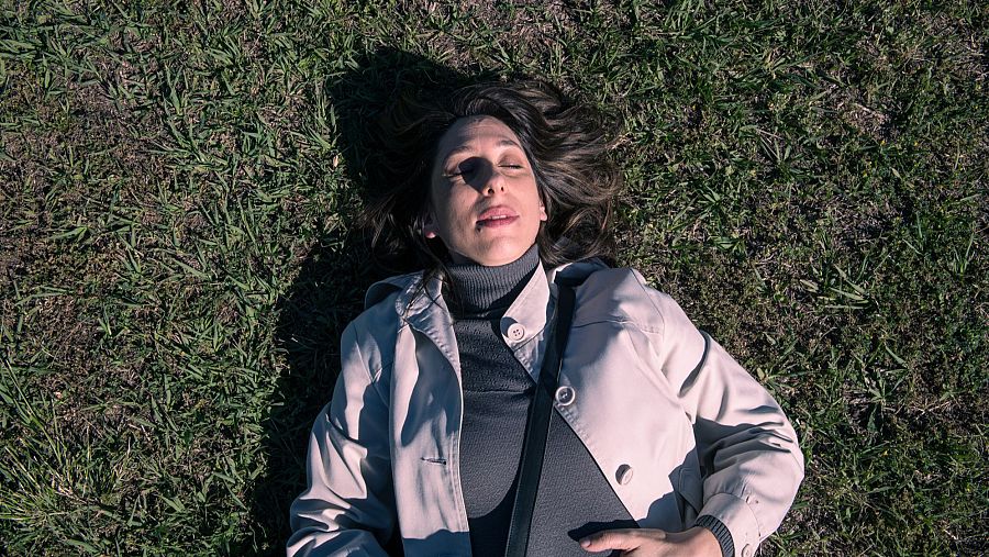 Mujer descansando boca arriba en césped, con gabardina beige, jersey gris y bolso negro. Cabello castaño, ojos cerrados, iluminada por el sol.