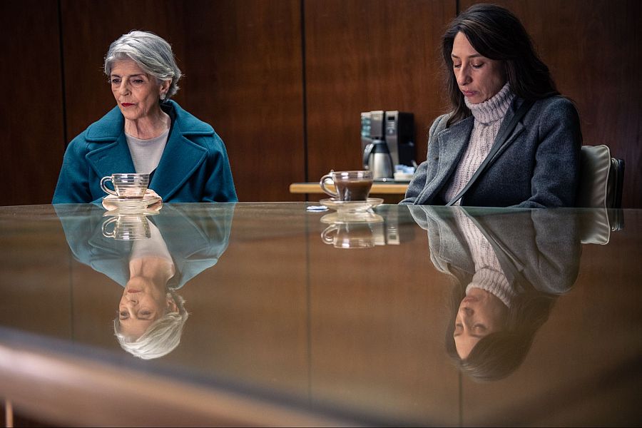 Dos mujeres, una con pelo corto y canoso, otra con pelo largo, conversan en una mesa de cristal.  Ambas parecen pensativas; la mesa refleja sus imágenes.