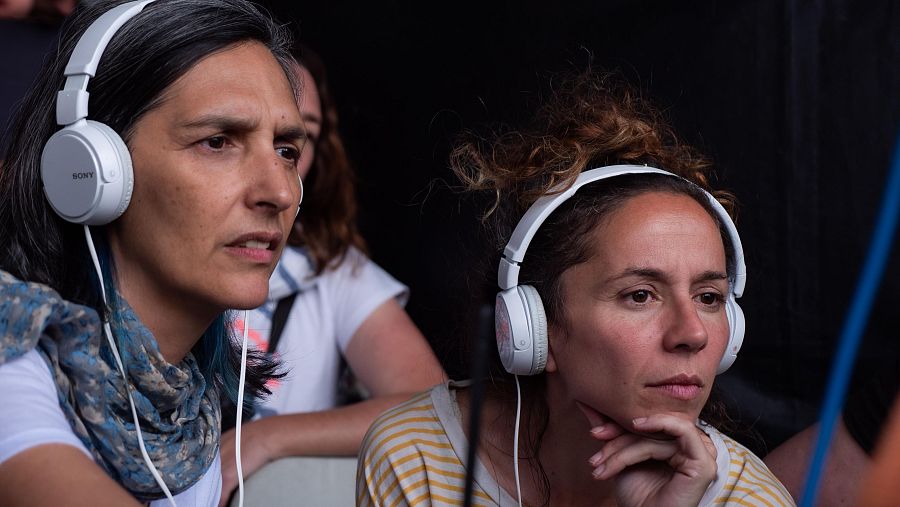 Dos mujeres con auriculares blancos, una con bufanda y otra con camiseta a rayas, muestran expresiones serias durante una grabación.