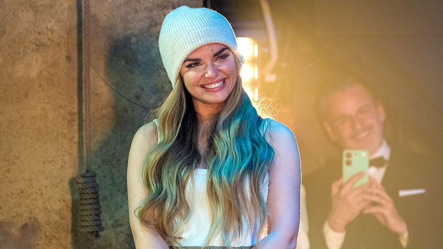 Cantante con gorro y vestido blanco, visiblemente emocionada, siendo grabada por un hombre con traje en un escenario de piedra.