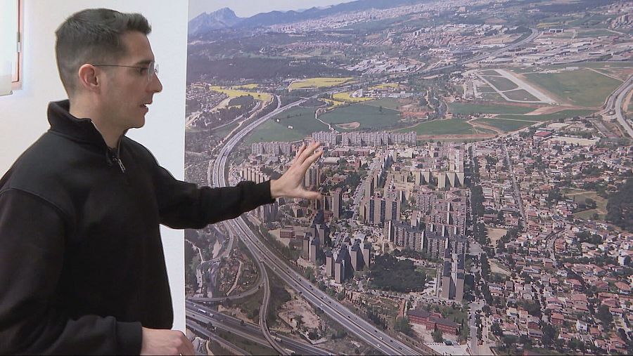 Alcalde de perfil, con gafas y chaqueta negra, señalando una fotografía aérea de gran escala que muestra edificios, carreteras y zonas verdes.