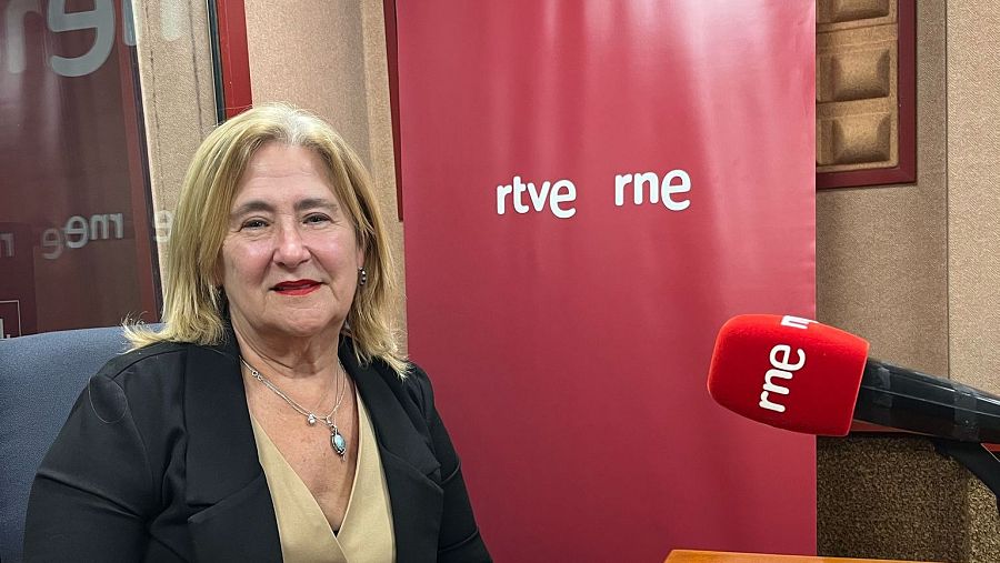 Mujer rubia con chaqueta negra en estudio de radio RNE, frente a micrófono 'rne' y panel con logo RTVE.  Posible programa cultural.