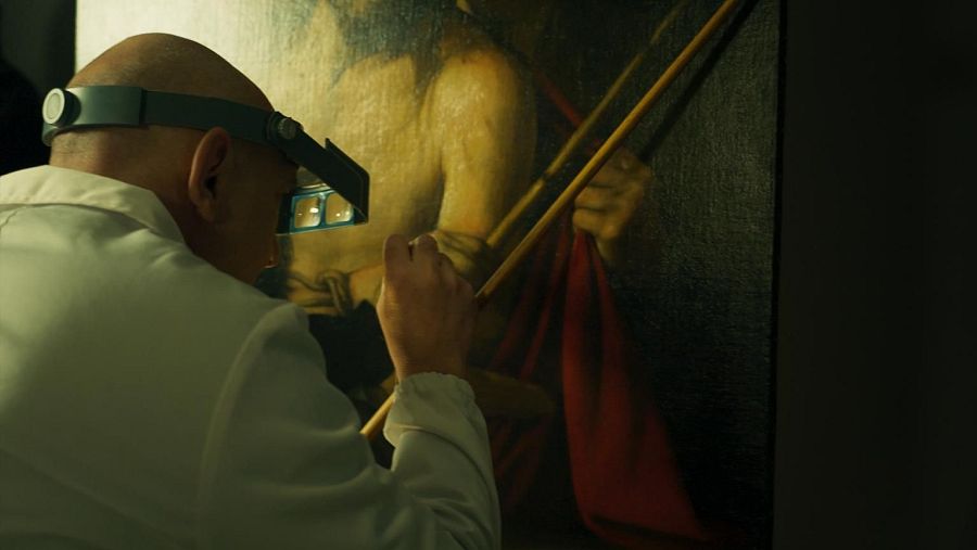 Hombre con bata blanca y gafas lupa examina pintura de figura masculina desnuda con instrumento fino.