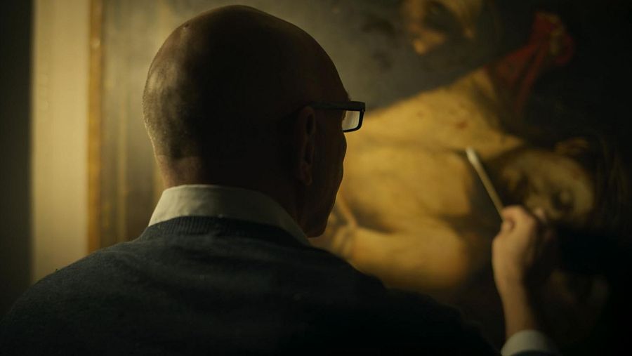 Restaurador examina con herramienta una pintura de tonos cálidos que muestra una figura reclinada, posiblemente masculina, con un paño en la cabeza.