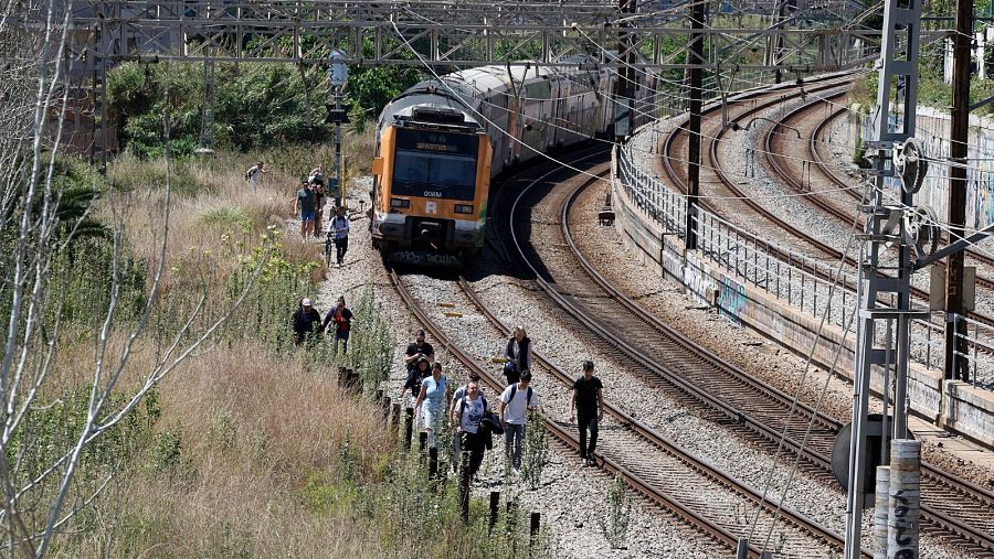 Tren en vía curva con personas caminando; vegetación seca y catenarias visibles.  Titular: 
