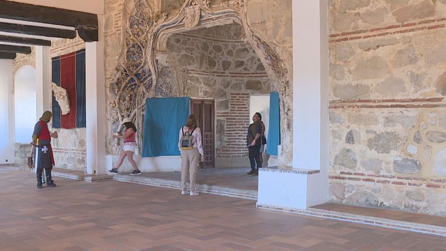 Amplio salón con suelo de baldosas y paredes de piedra; una arcada destaca como entrada a otra sala.  Una persona viste traje medieval, junto a otros visitantes.