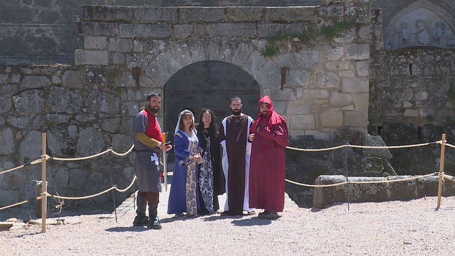 Cinco figurantes con trajes de época recrean un evento histórico frente a la entrada de un castillo de piedra. Una cuerda separa a los actores de los espectadores.